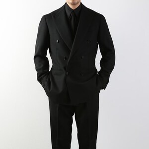 May include: A black double-breasted suit with a matching black shirt and tie. The suit has a classic design with a notched lapel and a tailored fit. The model is standing with their hands in their pockets.