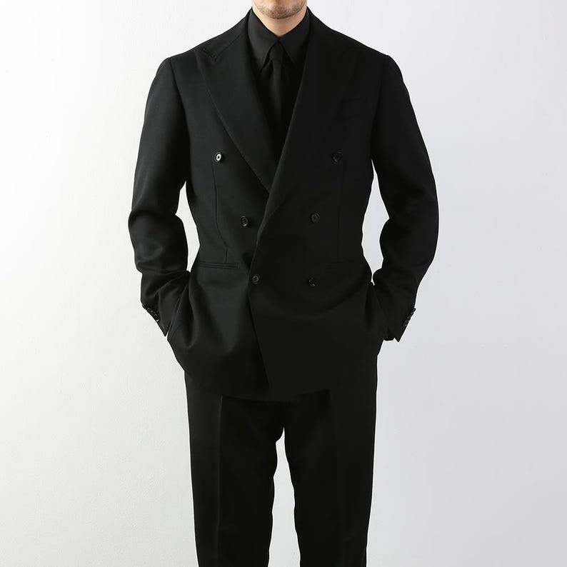 May include: A black double-breasted suit with a matching black shirt and tie. The suit has a classic design with a notched lapel and a tailored fit. The model is standing with their hands in their pockets.