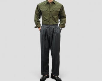 May include: An olive green button-down shirt with two chest pockets and dark gray pleated trousers. The outfit is completed with black loafers. The model is wearing a watch.