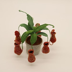 May include: A crocheted plant featuring green leaves and red-brown pitcher flowers. The plant is in a grey pot. The handmade plant is set against a neutral background. The crochet work is detailed and textured.