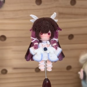 Può includere: Una bambola all'uncinetto fatta a mano con un design dettagliato. La bambola presenta capelli castani, dettagli viola e un vestito bianco. La bambola è montata su un'asta di metallo, mettendo in risalto l'artigianato e l'arte del pezzo.