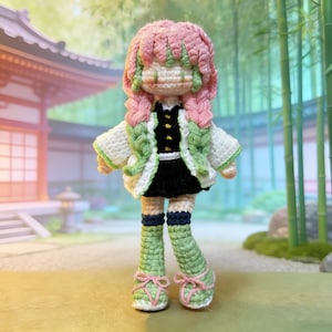May include: Crocheted doll with pink and green hair, wearing a black and white outfit with green accents. The doll has green and pink shoes with pink bows. The background features a Japanese-style building and bamboo.