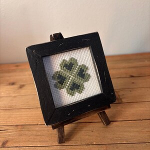 Peut inclure: Petite broderie au point de croix encadrée d'un trèfle à quatre feuilles vert. L'œuvre est présentée dans un cadre carré en bois noir, posée sur un petit chevalet en bois. Fond : mur clair et surface en bois.