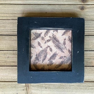 May include: A black wooden frame with a beige background and a pattern of brown feathers.
