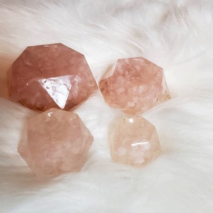 May include: Four pink, faceted, geometric crystal shapes with a white, cloudy, translucent appearance. The crystals are arranged on a white, fluffy surface.