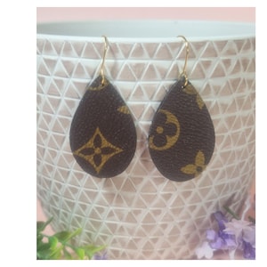 以下が含まれることがあります： 茶色の革のような外観の、しずく型のイヤリングのペア。イヤリングは金色のフックと、繰り返される金色の花柄が特徴です。イヤリングは、白いテクスチャード加工のベースの前に表示されています。