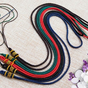 May include: A collection of colorful braided cords in red, green, black, and blue. Some cords have decorative gold and black accents. The cords are arranged on a neutral-toned fabric background.