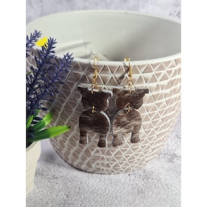 May include: A pair of dangle earrings featuring a brown and white cowhide pattern in the shape of a cow. The earrings are suspended from gold-colored hooks. The earrings are displayed in a white textured pot with a lavender plant.