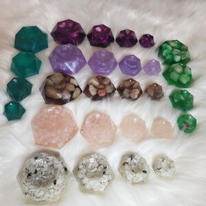 May include: A collection of 20 resin gemstone dishes in various colors and designs, including purple, green, and pink. The dishes are shaped like geometric gems and have different textures, including glitter, pebbles, and sand.