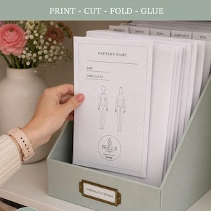 May include: A light green desktop file organizer containing white sewing pattern envelopes. The front envelope features a line drawing of a human figure, the text "PATTERN NAME," "SIZE," "DIFFICULTY," and the "BELLS" logo. A brass label holder reads "SEWING PATTERNS."