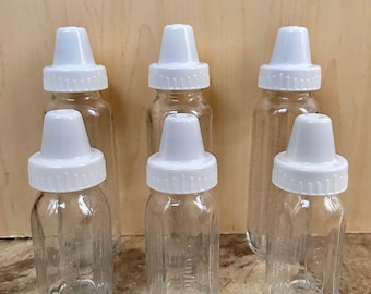 Op de afbeelding: Een set van zes doorzichtige glazen babyflesjes met witte plastic schroefdoppen. De flesjes zijn in twee rijen geplaatst, met drie grotere flessen achteraan en drie kleinere vooraan, allemaal voorzien van een geribbelde hals.