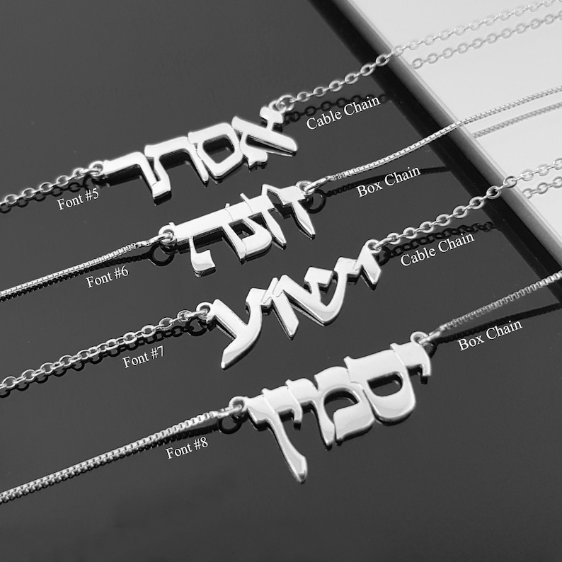 a collection of silver necklaces with Hebrew text engraved on them. The necklaces are arranged in a row, each with a different font and chain style.