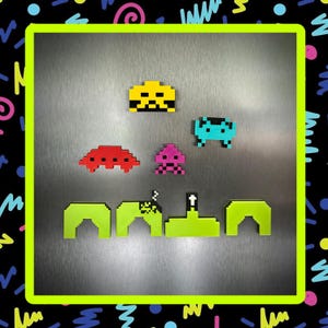 Op de afbeelding: Een verzameling gepixelde magneten in de stijl van het klassieke arcadespel, Space Invaders. De magneten zijn in verschillende kleuren, waaronder geel, rood, roze en blauwgroen. Het woord "GALAX" is in groene gepixelde letters geschreven.