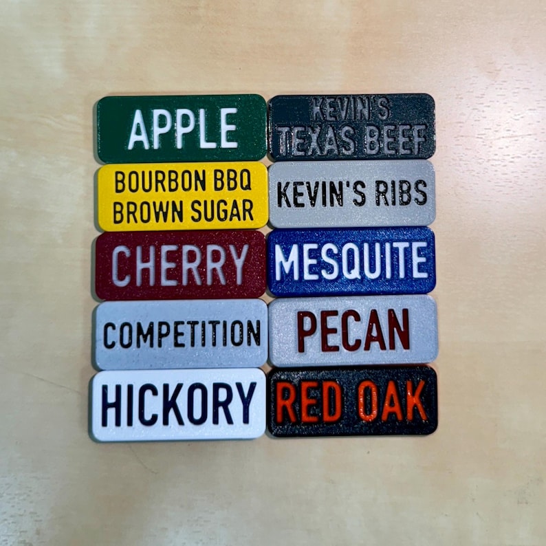 May include: Eight rectangular wooden signs with different wood smoking flavours printed on them. The signs are in various colours, including green, yellow, red, blue, and grey. The text on the signs includes: "Apple", "Bourbon BBQ Brown Sugar", "Cherry", "Competition", "Hickory", "Kevin's Texas Beef", "Kevin's Ribs", "Mesquite", "Pecan", and "Red Oak".