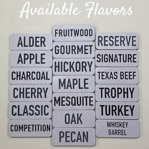 May include: A silver metal sign with the text "Available Flavors" at the top. Below the text are 16 rectangular silver metal signs with black text listing different wood flavours, including Alder, Apple, Charcoal, Cherry, Classic, Competition, Fruitwood, Gourmet, Hickory, Maple, Mesquite, Oak, Pecan, Reserve, Signature, Texas Beef, Trophy, Turkey, and Whiskey Barrel.