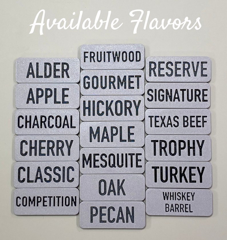May include: A silver metal sign with the text "Available Flavors" at the top. Below the text are 16 rectangular silver metal signs with black text listing different wood flavours, including Alder, Apple, Charcoal, Cherry, Classic, Competition, Fruitwood, Gourmet, Hickory, Maple, Mesquite, Oak, Pecan, Reserve, Signature, Texas Beef, Trophy, Turkey, and Whiskey Barrel.
