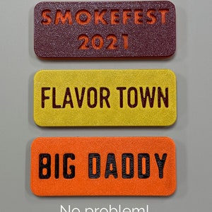 May include: Three rectangular tags with different text and colours. The top tag is maroon with orange text that says "SMOKEFEST 2021". The middle tag is yellow with black text that says "FLAVOR TOWN". The bottom tag is orange with black text that says "BIG DADDY".