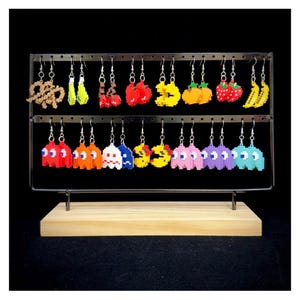 Puede incluir: Exhibición de pendientes pixelados con formas de pretzels, cerezas, fresas, plátanos y personajes de Pac-Man. Los pendientes están colocados en un expositor metálico de dos niveles con base de madera, sobre un fondo negro.