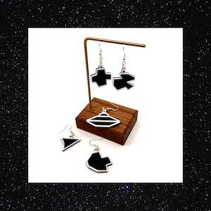 Puede incluir: Un conjunto de cuatro pendientes geométricos en blanco y negro en un expositor de madera. Los pendientes tienen forma de triángulo, OVNI, cruz y estrella.