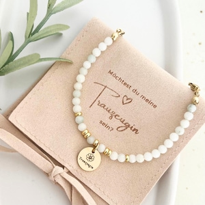 Könnte beinhalten: Ein zartes Armband mit weißen Perlen und goldenen Akzenten, mit einem runden Anhänger mit Blumenmuster und dem Wort "Trauzeugin". Das Armband liegt auf einem rosa Beutel mit deutschem Text.
