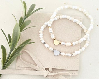 May include: Two white pearl bracelets with gold and silver heart charms. The bracelets are displayed on a beige fabric pouch with a drawstring.