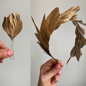 Op de afbeelding: Twee gouden veren haarclips, een is een enkele veer en de andere is een grotere groep veren. De verenclusters zijn bevestigd aan haarspeldjes.