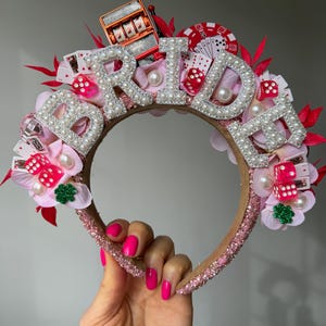 May include: A decorative headband with the word "BRIDE" spelled out in pearl-studded letters. The headband is adorned with pink flowers, dice, playing cards, a slot machine, and a casino chip. The base is brown with pink glitter.