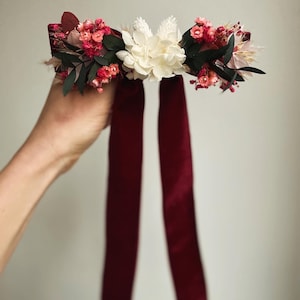 Puede incluir: Un lazo de terciopelo burdeos con un arreglo floral de flores secas rosas y blancas.