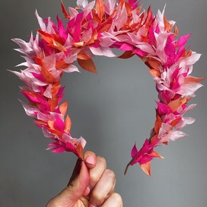 May include: A pink and orange floral crown made of paper leaves. The crown is held in a hand with pink nail polish.