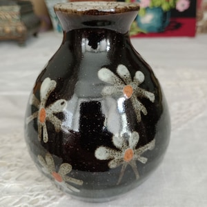 May include: A small, dark brown ceramic vase with a glossy finish. The vase is decorated with white and orange stylised flowers.