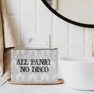May include: A white cosmetic bag with a black zipper and the text "ALL PANIC! NO DISCO" in black. The bag has a gray damask pattern and sits on a wooden tray. The background includes a white sink, mirror, and tiled wall.