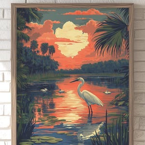 May include: A framed art print depicting a serene wetland scene. A white heron stands in the water, reflecting the sunset's orange and pink hues. Palm trees and lush greenery frame the water, with a vibrant sky filled with clouds.