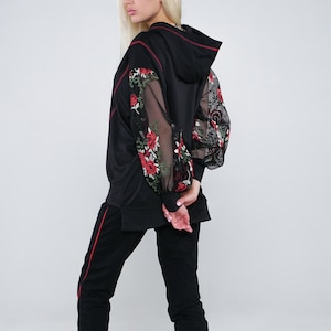May include: Black tracksuit with red accents. The hooded sweatshirt features mesh sleeves embroidered with red roses. The matching pants have a red stripe down the side. White and red sneakers complete the outfit.