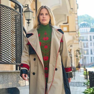 Puede incluir: Una mujer lleva una gabardina beige con ribetes rojos y un suéter con estampado floral verde y rojo. La gabardina tiene un detalle de cuero negro en la manga.