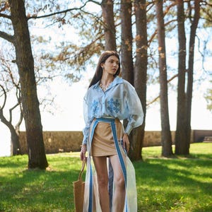 K&ouml;nnte beinhalten: Ein hellblaues, lang&auml;rmliges Oberteil mit Stickereien, kombiniert mit einem beigefarbenen Rock und einem transparenten &Uuml;berrock mit blauen Verzierungen. Das Outfit wird mit einer Korbtasche und Sandalen erg&auml;nzt. Die Szene spielt im Freien mit B&auml;umen und gr&uuml;nem Gras.