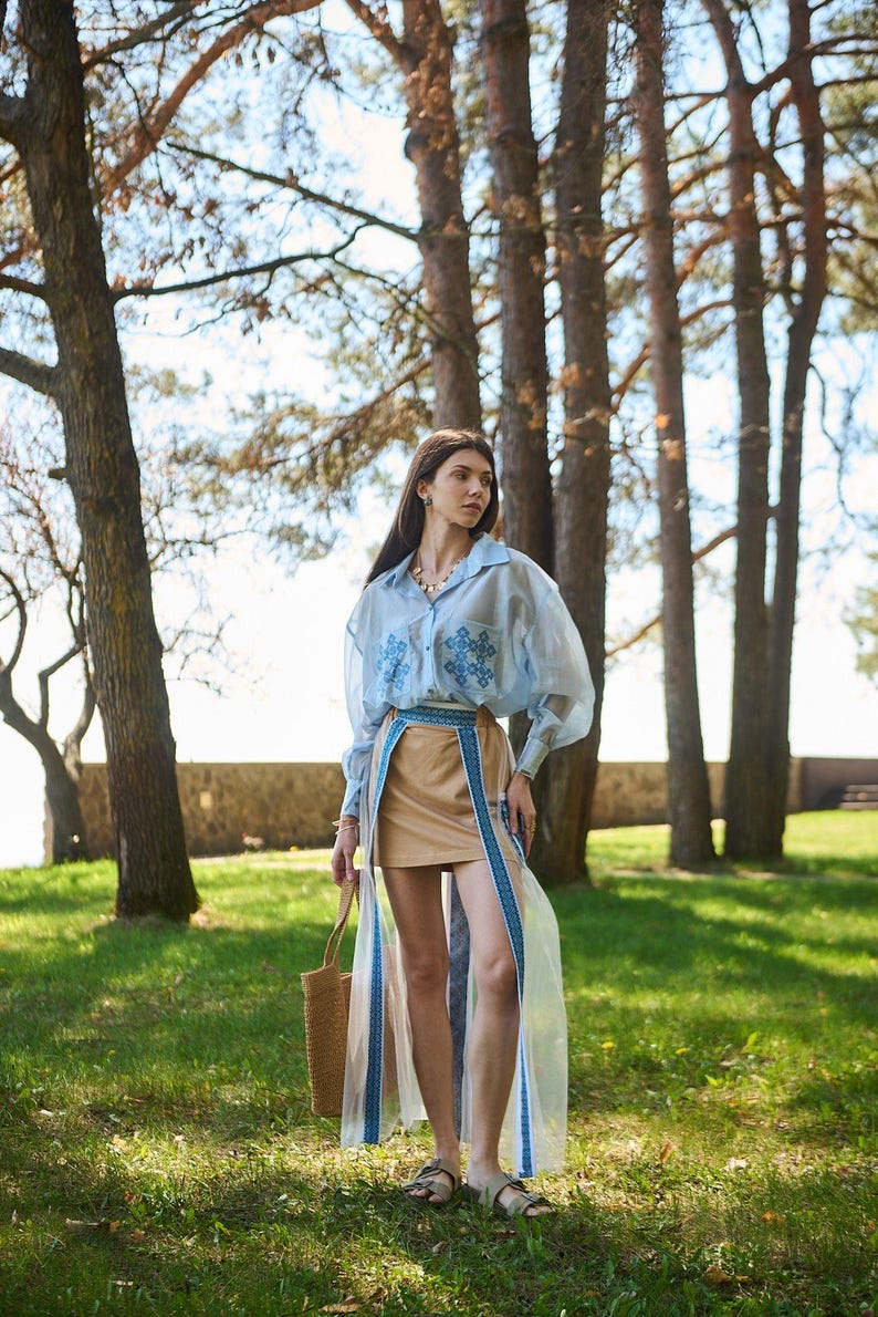 K&ouml;nnte beinhalten: Ein hellblaues, lang&auml;rmliges Oberteil mit Stickereien, kombiniert mit einem beigefarbenen Rock und einem transparenten &Uuml;berrock mit blauen Verzierungen. Das Outfit wird mit einer Korbtasche und Sandalen erg&auml;nzt. Die Szene spielt im Freien mit B&auml;umen und gr&uuml;nem Gras.