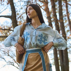 K&ouml;nnte beinhalten: Hellblaues Hemd mit Stickereien auf den Taschen und &Auml;rmeln, kombiniert mit einem beigefarbenen Rock mit blauem Stickbesatz. Eine Korbtasche wird &uuml;ber der Schulter getragen. Das Outfit befindet sich im Freien, mit B&auml;umen im Hintergrund.