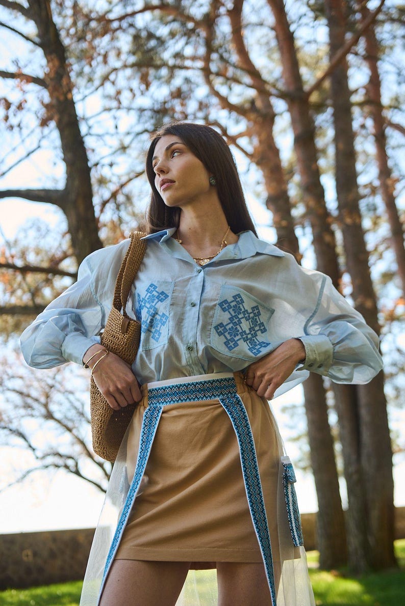 K&ouml;nnte beinhalten: Hellblaues Hemd mit Stickereien auf den Taschen und &Auml;rmeln, kombiniert mit einem beigefarbenen Rock mit blauem Stickbesatz. Eine Korbtasche wird &uuml;ber der Schulter getragen. Das Outfit befindet sich im Freien, mit B&auml;umen im Hintergrund.