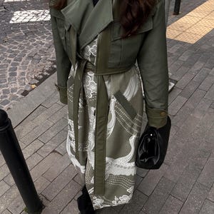 May include: An olive green trench coat with a matching belt and a patterned interior featuring white cranes. The coat is paired with black boots and a black handbag. The coat has a cropped jacket style over a longer dress.