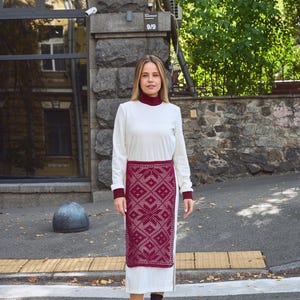 Peut inclure: Une femme portant une robe blanche à manches longues avec un panneau brodé bordeaux. Elle se tient sur un passage piétonnier devant un bâtiment en briques.