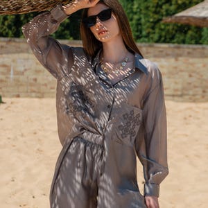May include: A gray silk button-down shirt and matching pants set. The shirt has a pocket with an embroidered design. The model is wearing black sunglasses and a gold necklace. The outfit is suitable for warm weather.