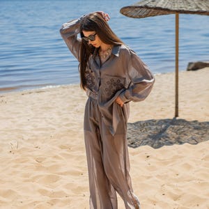May include: A light gray, two-piece outfit consisting of a button-down shirt and wide-leg pants. The shirt features embroidered detailing on the front. The outfit is worn on a sandy beach with the ocean in the background.
