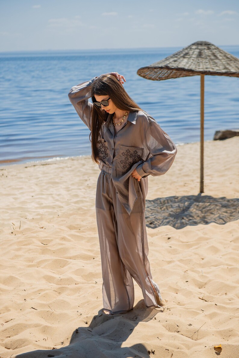 May include: A light gray, two-piece outfit consisting of a button-down shirt and wide-leg pants. The shirt features embroidered detailing on the front. The outfit is worn on a sandy beach with the ocean in the background.