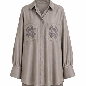 May include: A long-sleeved, button-down shirt in a muted taupe color. The shirt features two front pockets with embroidered geometric patterns in a darker shade of taupe. The sleeves have buttoned cuffs, and the shirt has a curved hemline.