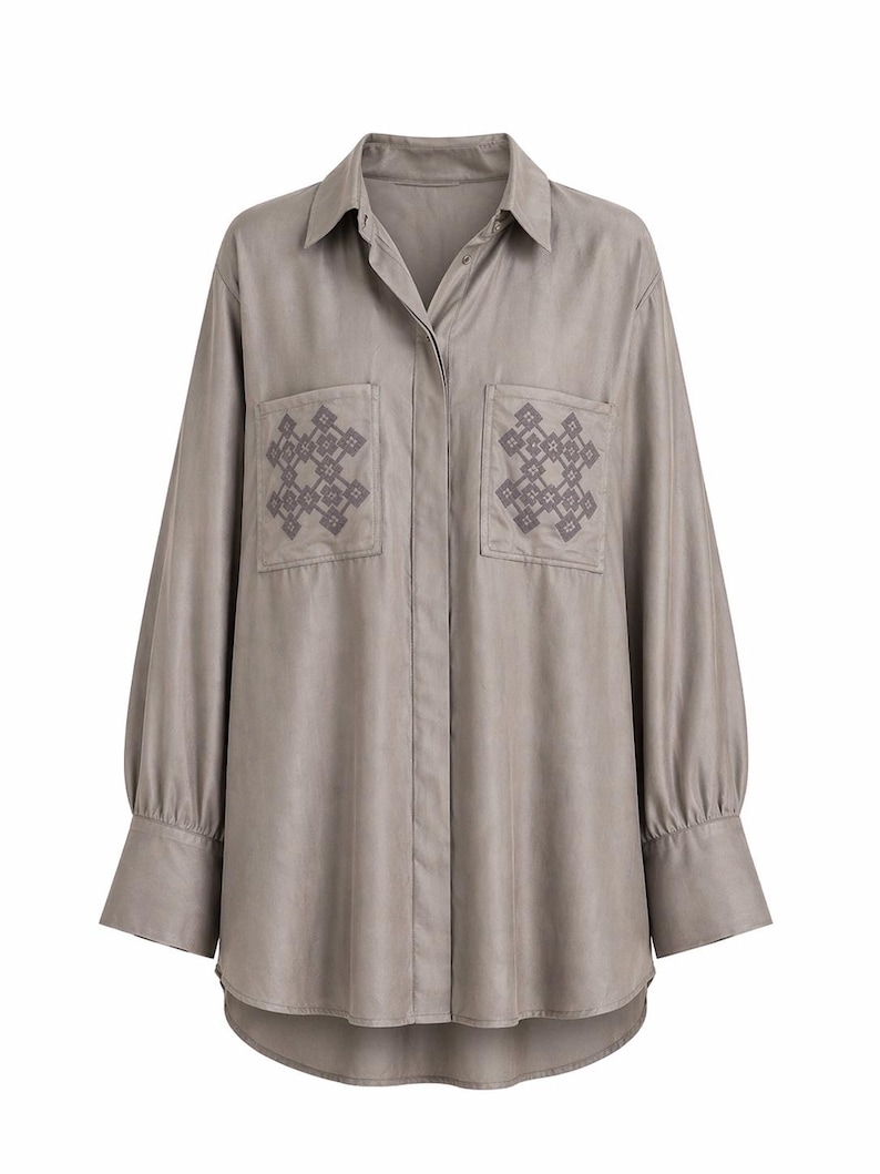 May include: A long-sleeved, button-down shirt in a muted taupe color. The shirt features two front pockets with embroidered geometric patterns in a darker shade of taupe. The sleeves have buttoned cuffs, and the shirt has a curved hemline.