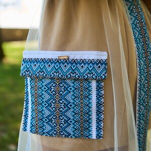 K&ouml;nnte beinhalten: Beiges Kleidungsst&uuml;ck mit einer blau-wei&szlig; gemusterten Tasche und Besatz. Die Tasche hat ein geometrisches Design in Blau-, Wei&szlig;- und Schwarzt&ouml;nen. Ein Holzakzent ist am oberen Rand der Tasche sichtbar. Das Kleidungsst&uuml;ck ist teilweise mit einem durchsichtigen, hellfarbigen Stoff bedeckt.