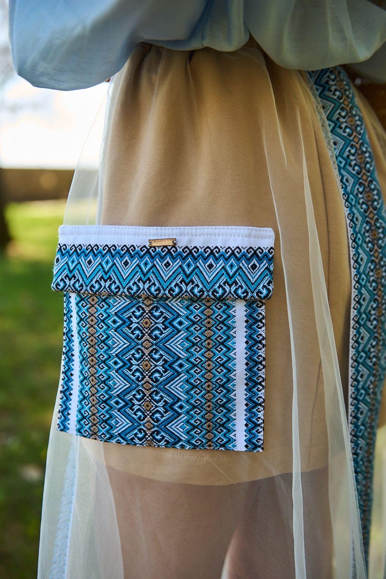 K&ouml;nnte beinhalten: Beiges Kleidungsst&uuml;ck mit einer blau-wei&szlig; gemusterten Tasche und Besatz. Die Tasche hat ein geometrisches Design in Blau-, Wei&szlig;- und Schwarzt&ouml;nen. Ein Holzakzent ist am oberen Rand der Tasche sichtbar. Das Kleidungsst&uuml;ck ist teilweise mit einem durchsichtigen, hellfarbigen Stoff bedeckt.