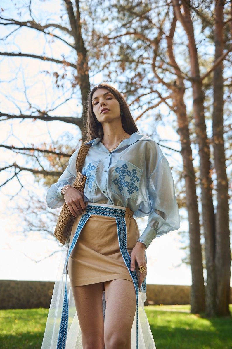 K&ouml;nnte beinhalten: Ein hellblaues, durchsichtiges Hemd mit Kn&ouml;pfen und gestickten blauen geometrischen Designs auf den Taschen. Das Outfit umfasst einen beigefarbenen Rock mit einem blau-wei&szlig; gemusterten G&uuml;rtel und eine gewebte Tasche. Ein durchsichtiger wei&szlig;er Rock-Overlay ist sichtbar.