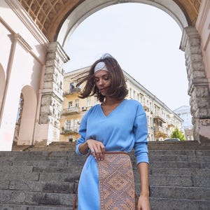 Peut inclure: Une femme portant un pull bleu clair et une jupe en crochet marron se tient sur un escalier en pierre. Elle porte un bandeau blanc et ses cheveux sont balayés par le vent. Elle tient un sac cabas bleu clair.