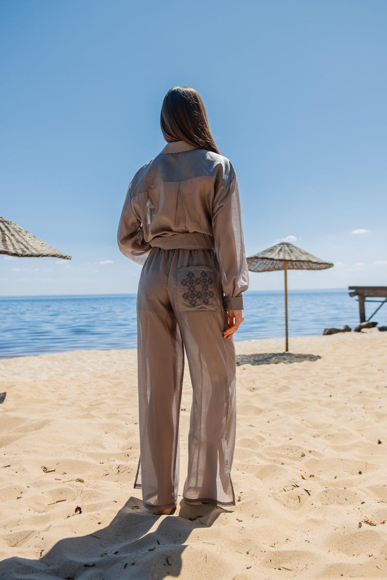 May include: A light brown, two-piece outfit on a sandy beach. The outfit includes a long-sleeved button-up shirt and wide-leg pants. The pants have a decorative design on the back pocket. The background features the ocean and a blue sky.